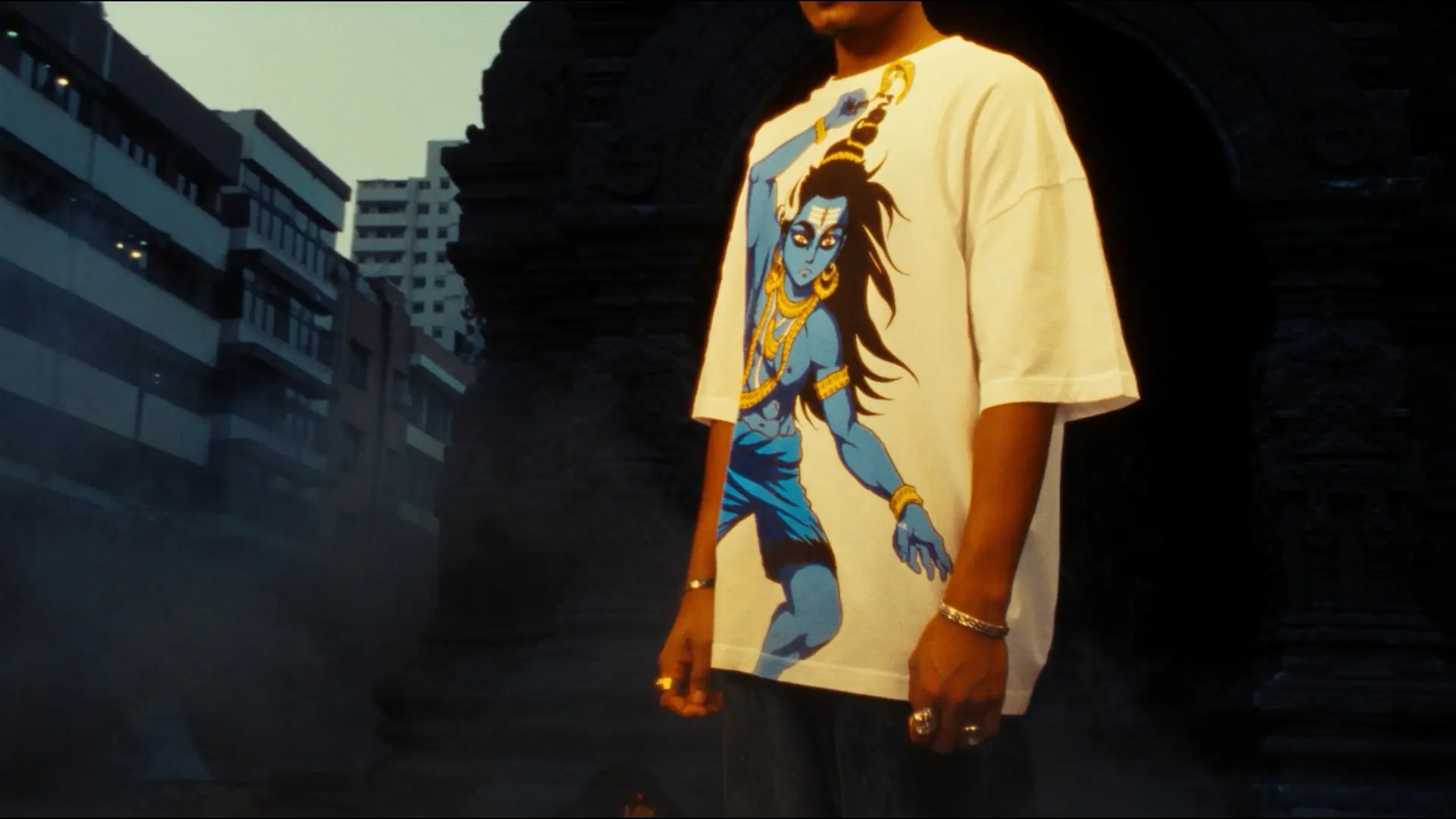 Young Indian man wearing premium oversized white tshirt with Lord Shiva rendered in anime illustration style, sacred temple archway backdrop, golden hour lighting